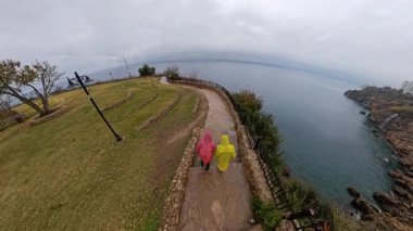 Parlak Yağmurluklu Bir Erkek ve Kadın Uçurum Yamacında Yürüyor, Denize Bakıyor, Şehir Skyline 'ı ve Arka plandaki Engebeli Sahil Çizgisi ile