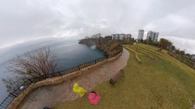 Pembe Yağmurluk Giyen Bir Adam ve Sarı Yağmurluk Giyen Bir Kadın Denize Bakan Taş Yol 'da Yürüyorlar Şehir Binaları ve Uçurumları Bulutlu Bir Gün Manzarasını Çerçeveliyor