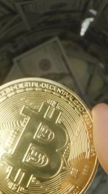 Dikey video. 100 Dolarlık banknotlarla dolu bir cam kavanoza düşerken Altın Bitcoin 'in Orta Havada Yakından Görünüşü, Atışın Hareketini Yakalıyor. Dijital ve Fiziksel Zenginliğin Bütünleşmesi