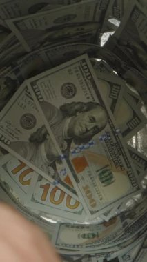 Dikey video. Yüz dolarlık banknotlarla dolu bir kavanozdan Altın Bitcoin 'i çıkaran Parmakların Yakın Çekim Görüntüsü, Sikke ve İç Kesim' e odaklanıyor. Dijital Zenginlik Çekimi Konsepti