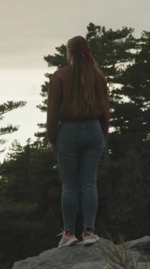 Dikey video. Kayalık bir uçurumun kenarında duran genç bir kadın, sırtı kameraya dönük, sık bir orman ve altın gün batımına bakıyor, uzun saçları kırmızı bir kurdeleyle bağlanmış. Huzur ve Yansıma