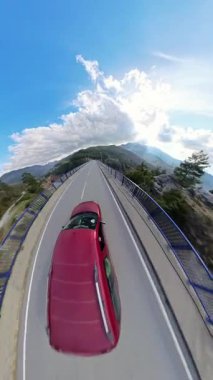 Dikey video. Köprüden geçen kırmızı araba gölün manzaralı manzarası orman tepeleri ve dinamik bulutlarla dolu parlak bir gökyüzünün altındaki dağlar. Yükseltilmiş Sahne Sürücüsü