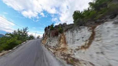 Keskin kıvrımları kayalık yamaçlar ve seyrek yeşillikler tarafından çevrili, açık mavi bir gökyüzünün altında dağınık bulutlarla uzanan virajlı toprak bir yol. Dağlık Yol