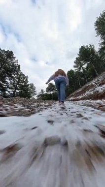 Dikey video. Çam ağaçları ve parçalı bulutlu bir gökyüzü ile çevrili kar tozlu kayalık bir yolda yokuş yukarı yürüyen bir kadının arka görüntüsü. Dağ Yolu