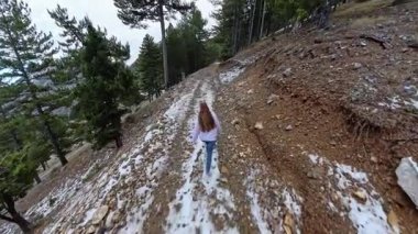 Geniş görüşlü, uzun saçlı bir kadın, parlak bir gökyüzü altında, çam ağaçları ve uzak dağ manzaralarıyla çevrili bir ormanda, tozlu kayalık bir patikada yürüyor. Huzurlu Kış Yürüyüşü
