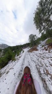 Dikey video. Karla kaplı patika çam ağaçları ve kayalık araziyle ormanlık bir yamaçta kıvrılarak bulutlu bir gökyüzüne ve uzak dağlara doğru ilerliyor. Sakin Kış Yolu