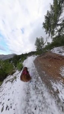 Dikey video. Ağaçlarla çevrili, açık bir gökyüzü ve uzak dağların zirveleriyle karla kaplı patikada yürüyen kırmızı ekose kurdeleli bir kadının yukarıdan görünüşü. Huzurlu Kış Gezintisi