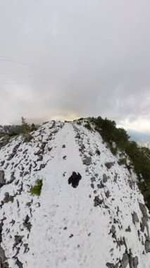 Dikey video. Küçük gezegen etkisi, engebeli arazi, dağınık çam ağaçları ve dramatik bulutlu gökyüzü ile çevrili karlı bir dağ sırtında yürüyen bir adamı yakalıyor. Gerçeküstü Kış Keşfi
