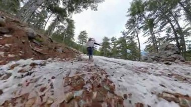 Bulutlu bir gökyüzünün altında, yüksek bir perspektiften yakalanmış, uzun çam ağaçlarıyla çevrili bir ormanda, karlı bir yamaçtan aşağıya koşan bir kadın. Kış Macerası