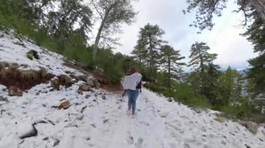 Bulutlu bir gökyüzü ve uzak dağ manzarası olan yamaçtaki yüksek çam ağaçlarıyla çevrili karla kaplı bir orman patikasında koşan bir kadın. Kış Ormanı Koşusu