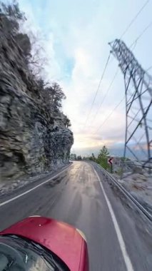 Dikey video. Kayalık uçurumlara oyulmuş sarmal bir dağ yolunun perspektif görüntüsü, kenarlarında çam ağaçları ve yumuşak akşam ışığı ile aydınlanan bulutlu bir gökyüzü. Manzaralı Dağ Sürüşü