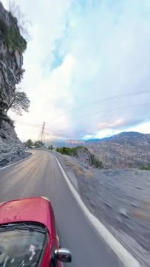 Dikey video. Yukarısında dramatik bulutlar ve elektrik hatları olan kayalık kayalıklar ve çam ağaçlarıyla çevrili ıslak, dolambaçlı bir dağ yolunda giden kırmızı bir arabanın alçak açılı görüntüsü. Maceracı Sürüş