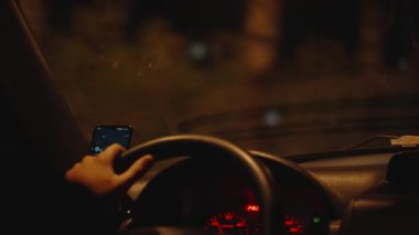 Sürücü Eli Direksiyonu Smartphone Navigasyon Ekranı Görünür, Aydınlatılmış Kontrol Paneli Işıkları ve Bulanık Gece Görüşü Ön camdan. Gece Sürücüsü Odaklanma