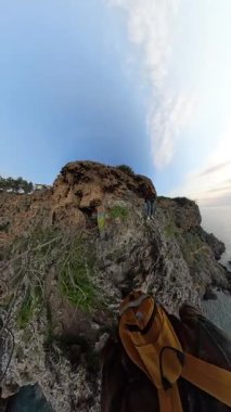 Dikey video. Bir çift kadın ve erkek gezgin dikkatle denize doğru kayalık bir uçurumdan iniyor. Etrafı bitki örtüsüyle çevrili ve 360 derecelik bir manzarayla çevrilmiş. Keşif ve Macera