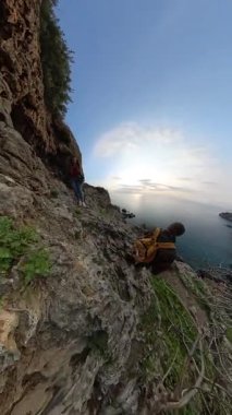 Dikey video. Bir erkek ve kadın gezgin, engebeli arazi ve bitki örtüsüyle çevrili engebeli bir kayalık yamaçtan denize doğru yol alıyor. Kıyı Keşfi