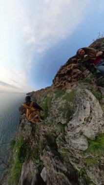 Dikey video. Bir Kadın ve Erkek Gezgin Denize Doğru Kayalık Kayalıklarda Geziniyor, Etrafı Bitkilerle çevrili ve 360 derecelik bir Etkiyle Ele Geçirilmiş. Macera ve Keşif