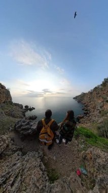 Dikey video. Günbatımında Okyanusa Bakan Kayalık Kayalıklarda Birlikte Duran Bir Kadın ve Erkek Gezgin, Sahneyi Çerçeveleyen Kayalıklarla ve Yukarıda Uçan Kuşla. Yolculuğa yansıyan