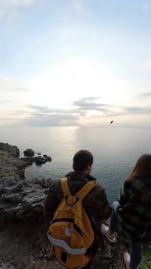 Dikey video. Bir Erkek ve Kadın Gezgin Okyanusa Bakan Uçan Kuş ve Uzakta Güneş Batan Uçan Bir Uçurumda Birlikte Duruyor. Serene Sahil Yansıması