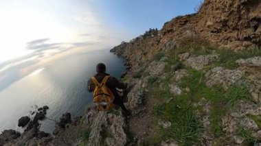 Sarı sırt çantalı bir erkek gezgin okyanusa doğru kayalık bir tepeden aşağı dikkatle yürüyor, engebeli arazi ve bitki örtüsüyle çevrili. Kıyı Seyahati