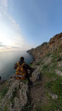 Dikey video. Sarı sırt çantalı bir erkek gezgin okyanusa doğru kayalık bir tepeden aşağı dikkatle yürüyor, engebeli arazi ve bitki örtüsüyle çevrili. Kıyı Seyahati