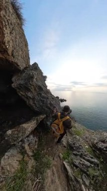 Dikey video. Altında Yürüyen Bir Kadın Gezgin Engebeli Uçurumlar ve Okyanusa Bakan Tepelerle çevriliyken Erkek Bir Gezgin Kayalık Yoldan Aşağı Kayıyor. Manzaralı Kıyı Düşüşü