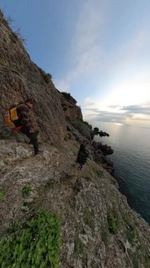 Dikey video. Bir Kadın ve Erkek Gezgin Dik Kayalık Kayalık Yol 'da Yürüyor, Etrafı Okyanusla çevrili ve Arka planda Güneş Batıyor. Sahne Kıyı Yolculuğu