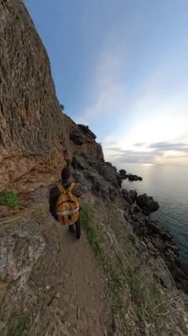 Dikey video. Bir Erkek ve Kadın Gezgin Rocky Cliffside Patikası boyunca yürürken etrafını bitki örtüsü, okyanus ve arka planda batan güneş sarar. Kıyı Keşfi