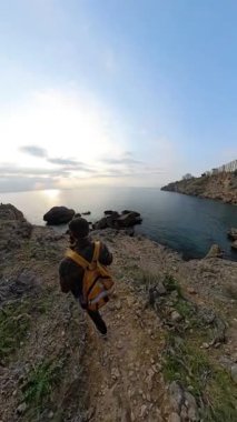 Dikey video. Rocky Cliffs ve Arkaplanda Batan Güneş 'le birlikte, Erkek ve Kadın Bir Gezgin Engebeli Kıyı Yolu' ndan Okyanusa Gidiyor. Sahne Kıyı Keşfi