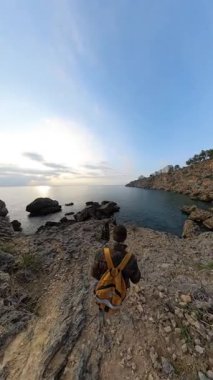 Dikey video. Bir Erkek ve Kadın Gezgin Kayalık Kayalıklar, Sakin Okyanus ve Uzakta Batan Güneşin Işıltısı ile Kayalık Yamaçtan Aşağı Kayalığa Yürüyor. Kıyı Yolculuğu