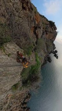 Dikey video. Bir Erkek Gezgin, Okyanusun Altında ve Geniş Açı Ufacık Gezegen Perspektifi ile Bir Kayalık Yol Düşürürken Bir Kadın Gezgine Yardım Eder. Destekleyici Kıyı Macerası