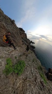 Dikey video. Bir Erkek Gezgin Kayalıklarda Dar Kayalık Yol 'da dikkatle Geziniyor, Bitki örtüsü ve Okyanus Altında, Benzersiz Küçük Gezegen Perspektifinde Yakalandı. Macera İnişi