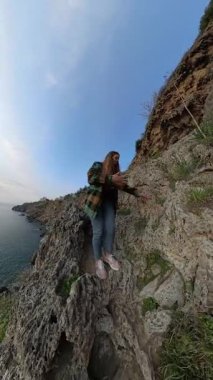 Dikey video. Dik Kayalık Kayalık Kayalıklardan Dikkatlice İnen Bir Dişi Gezgin, Çevresi Bitki örtüsü ve Okyanusa Bakış Alıyor. Solo Sahil Macerası