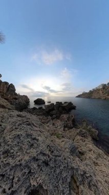 Dikey video. Panorama Bir Erkek ve Kadın Gezgin Okyanus kenarındaki Jagged Kayalıklarda Otururken, Kayalıklar Uzağa Uzanıyor ve Sakin Su Bulutlu Gökyüzünü Yansıtıyor. Sahne Kıyı Dinlenme