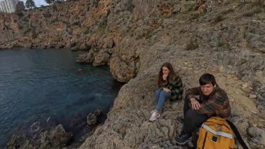 Uçurumun kenarındaki Jagged Kayalıklarda oturan bir Erkek ve Kadın Gezgin Bulutlu Gökyüzünün Ardındaki Gün Batımını izlerken Sakin Mavi Okyanusu seyrediyor. Huzurlu Kıyı Anı