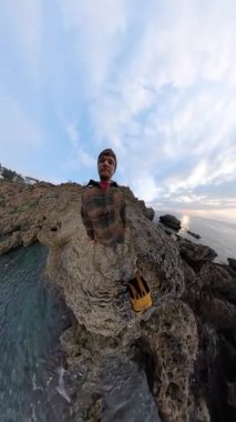 Dikey video. Okyanus kıyısındaki Jagged Rocks 'ta Sırt Sırtında Yürüyen Bir Adam, Yukarı Kaldırıyor ve Omuzlarına Koyuyor, Üçüncü Perspektiften Çekiyor. Engebeli Kıyı Sahnesi