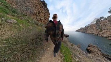 Uçurumun kenarındaki Dar Kayalık Yol 'da Yürüyen Bir Adam, Etrafı Bitkilerle çevrili ve Bulutlu Bir Gökyüzünün Altındaki Sakin Okyanusa Bakıyor. Engebeli Sahil Yolculuğu