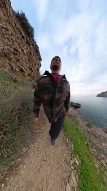 Dikey video. Uçurumun kenarındaki Dar Kayalık Yol 'da Yürüyen Bir Adam, Etrafı Bitkilerle çevrili ve Bulutlu Bir Gökyüzünün Altındaki Sakin Okyanusa Bakıyor. Engebeli Sahil Yolculuğu