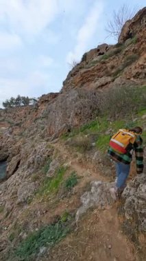 Dikey video. Sarı Sırt çantalı Bir Kadın Dar Kayalık Yol boyunca Dar Kayalıklarda Yürüyerek Aşağı Okyanusa ve Engebeli Araziye Bakar. Kıyı yürüyüşü macerası