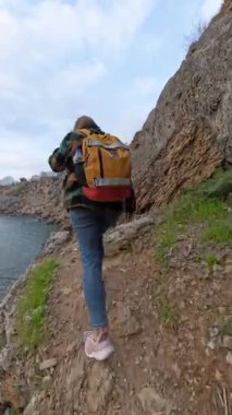 Dikey video. Sırt çantalı bir kadın uçurum kenarındaki sarp kayalıklara tırmanıyor, etrafını bitkiler sarmış ve aşağıdaki geniş okyanusa tepeden bakıyor. Kıyı Keşfi