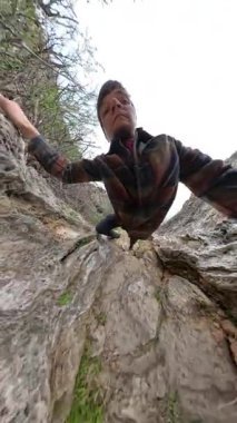 Dikey video. Tırtıklı kayalara tırmanan bir adam eliyle bir uçurumun kenarına tutunarak okyanusun altında, yukarı doğru bir açıdan yakalanır. Sert Kıyı Yükselişi