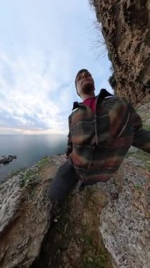 Dikey video. Ekose gömlekli bir adam uçurumun kenarındaki engebeli kayalık arazide tırmanıyor, azimle yukarı bakıyor, düşük açılı küresel panorama perspektifinden çekiliyor. Macera