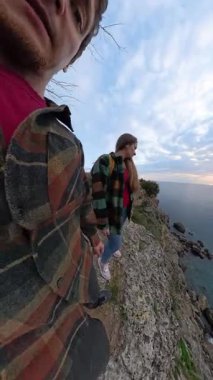 Dikey video. Ekose ceketli bir adam, uçurumun kenarında, denize bakan bir kadınla duruyor ve sarı sırt çantasıyla Bulutlu Gökyüzüne karşı çarpık küresel bir perspektifte yakalandı. Manzaralı Uçurum Macerası