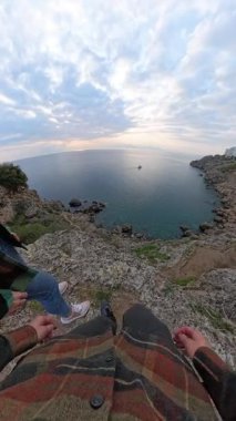 Dikey video. Ekose ceketli bir kadın, Rocky Cliff 'in üzerinde durmuş geniş bir sahil manzarasıyla denize bakıyor ve Güneş' i canlı gökyüzü ile küresel bir perspektiften yakalıyor. Kıyı Keşfi