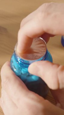 Dikey video. Mavi Plastik Kutuyu Haplarla Açan, Foil Mührü Kaldıran, Tahta Masa Yüzeyini Aydınlatan Yakın Çekimde, Sıcak Doğal Işıkta Yakalandı. Sağlık ve İlaç Konsepti