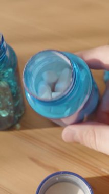 Dikey video. Beyaz ve Mavi Kapsüllerle Dolu Mavi Plastik Şişe, Arkaplandaki İki Açık Şişe ile Sıcak Doğal Işık Altındaki Ahşap Masada. Sağlık ve İlaç Örgütü Konsepti