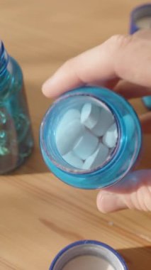 Dikey video. Beyaz Oval Tabletleri Mavi Şişe 'den Palmiye' ye döken Eller, Arka planda Diğer Şişelerle birlikte, Sıcak Doğal Işıkta Bir Ahşap Masada Yakından Çekildi. Sağlık ve Tıp Konsepti
