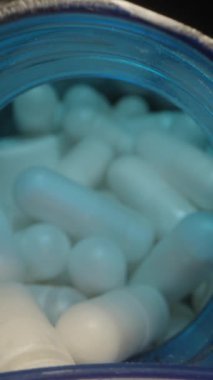 Dikey video. Beyaz Kapsüller Mavi Şişe 'den Lid' e dökülüyor, Keskin Macro Focus 'ta Karanlık Bir Arkaplana Karşı Aydınlatma ile yakalandılar. Sağlık ve Sağlık Hizmetleri