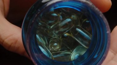 Mavi Şişe 'den Altın Şeffaf Jelatin Kapsülleri Döken Eller, Parlak Dokularını ve Yakın Görüşte Canlı Renkleri Vurguluyor. Sağlıklı Yaşam