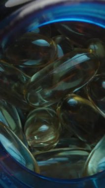 Dikey video. Mavi Şişe 'den Altın Şeffaf Jelatin Kapsülleri Döken Eller, Parlak Dokularını ve Yakın Görüşte Canlı Renkleri Vurguluyor. Sağlıklı Yaşam