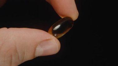Tek bir Altın Şeffaf Omega 3 Kapsülü 'nü Tutan Bir El Siyah Arkaplan Yedeklemesine Karşı Pürüzsüz Dokusunu ve Parlak Bitişini Vurguluyor